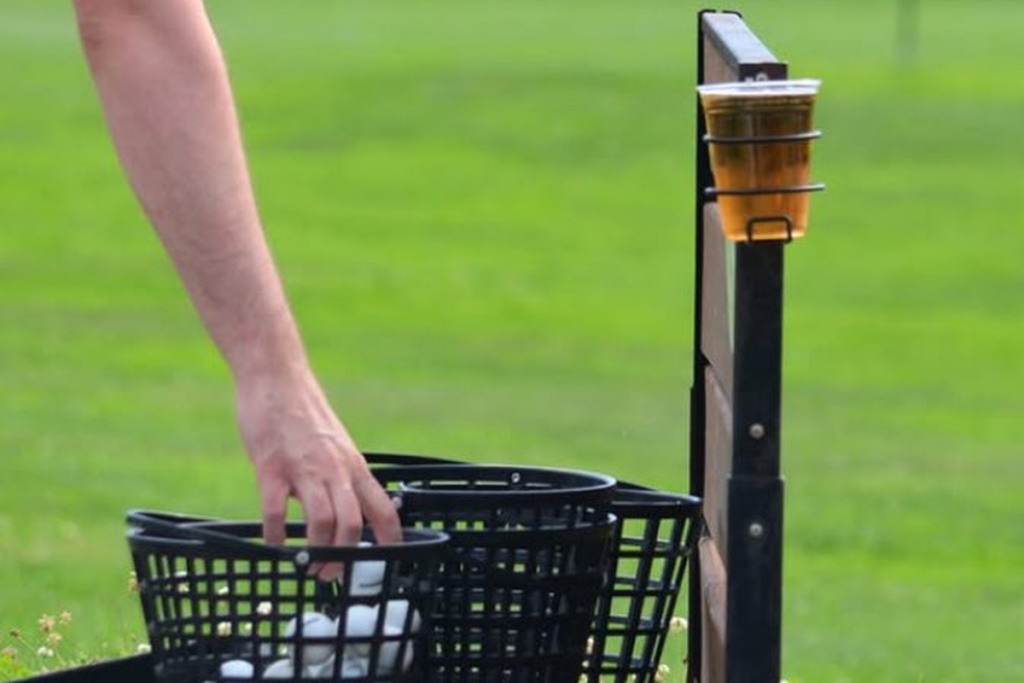 Beer resting in a driving range divider cup holder while a golfer grabs range balls